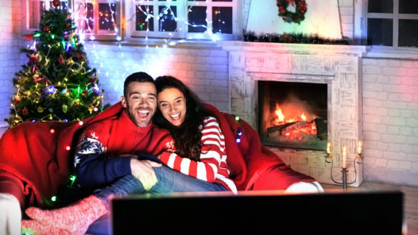 Smiling young couple having fun at home, watching TV together and laughing. Both wears warm clothes and sitting on sofa. On background lights on decorated christmas tree, windows and burning fireplace, steel candlestick on ground. Back of TV set on foreground.